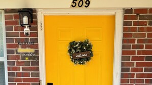 A bright yellow door with a welcome wreath, a number 509 sign above, and a security camera on the brick wall. - Sunshine Paradise - Elegant 2-bedroom unit in charming Orange Park, FL (Orange Park)