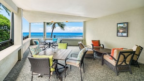 A covered outdoor seating area with a view of the ocean, featuring wicker furniture and a wooden table. - Banyan Tree - Kona Beach Properties (Kailua-Kona)