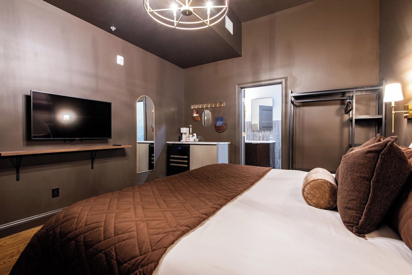 A hotel room with a large bed, a flat-screen TV, a vanity with a mirror, and a modern ceiling light fixture.