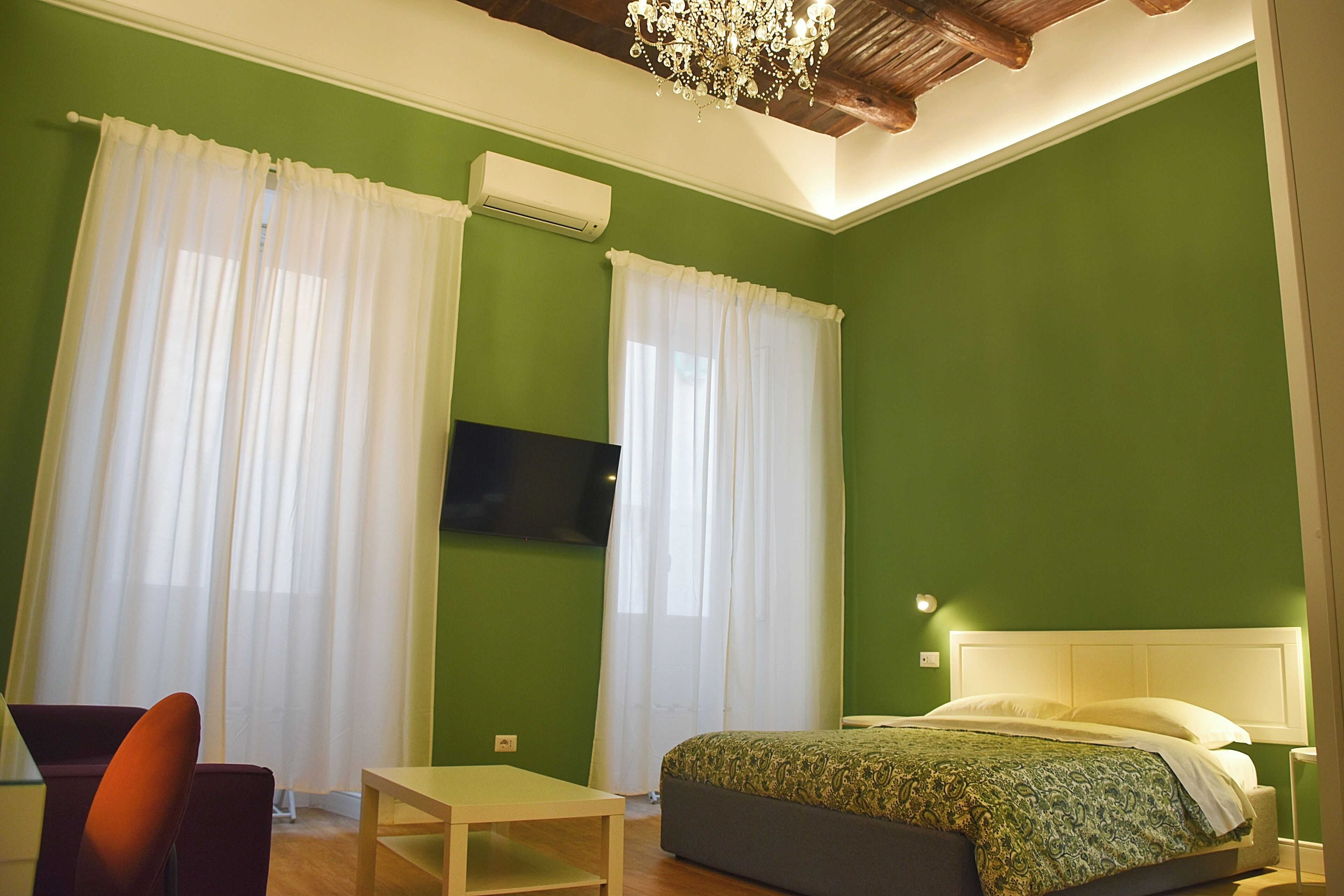 Un dormitorio con paredes verdes, una cama con ropa de cama estampada, un televisor montado en la pared y una araña.