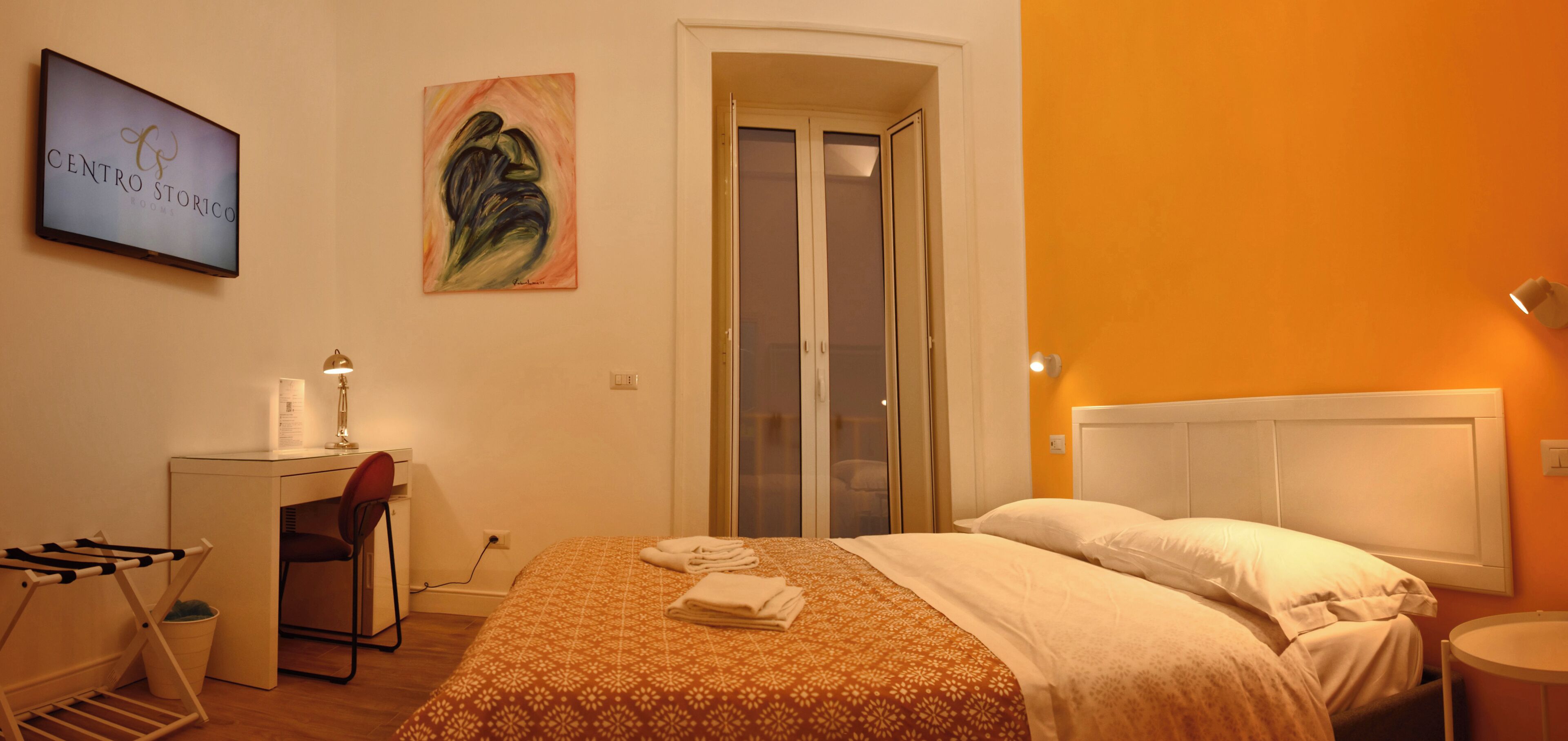 Een hotelkamer met een bed, een bureau, een stoel, een aan de muur bevestigde televisie en een deur naar een balkon.