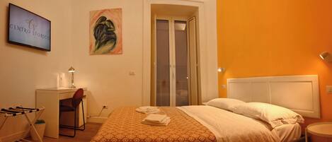 Habitación de hotel con cama, escritorio, silla, televisión montada en la pared y puerta que da a un balcón.