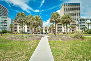 A pathway leading to a building complex with multiple floors and balconies, surrounded by palm trees and greenery. - Two bedroom steps from the beach fully updated! (Myrtle Beach)