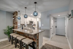 A modern kitchen with a central island, bar stools, hanging pendant lights, and a wooden accent wall. - Two bedroom steps from the beach fully updated! (Myrtle Beach)