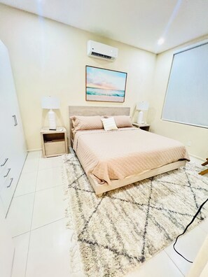 A bedroom with a bed, bedside tables, an air conditioner, a painting, and a view of the outdoors. - This gated community offer a Tranquil 1-bedroom condo with pool & 5 min to beach (Christ Church)