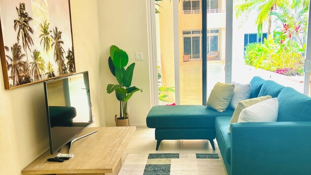 A modern living room with a sofa, a coffee table, a TV, and a large window with a view of a garden. - This gated community offer a Tranquil 1-bedroom condo with pool & 5 min to beach (Christ Church)