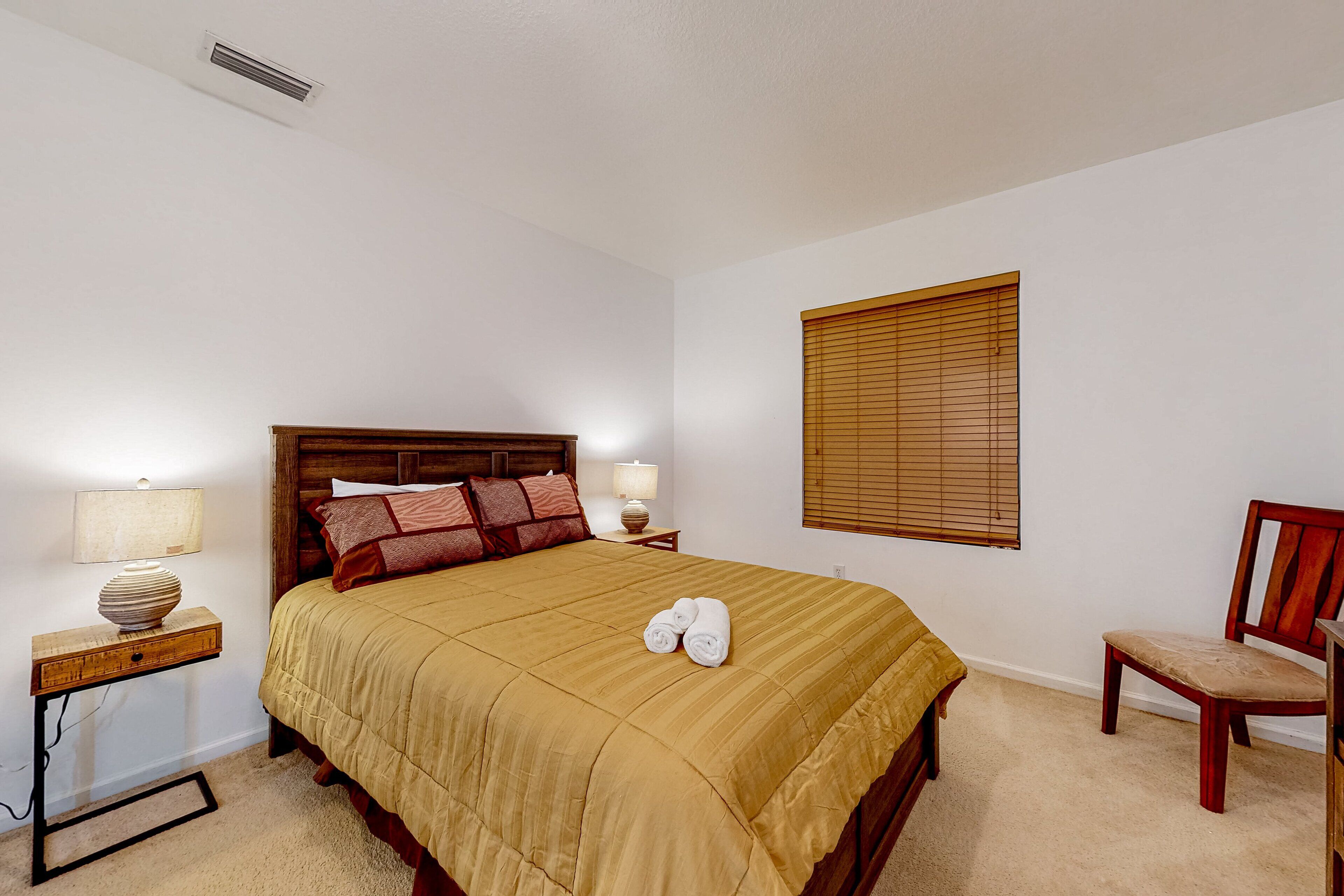 A neatly made bed with a beige bedspread, two bedside lamps, a wooden nightstand, a wooden chair, and a window with blinds.