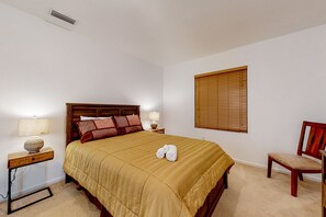 A neatly made bed with a beige bedspread, two bedside lamps, a wooden nightstand, a wooden chair, and a window with blinds. - Palm Bay Cove (Palm Bay)