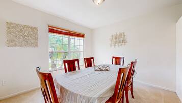 A dining room with a large table, six chairs, a window with red blinds, and two wall art pieces.
