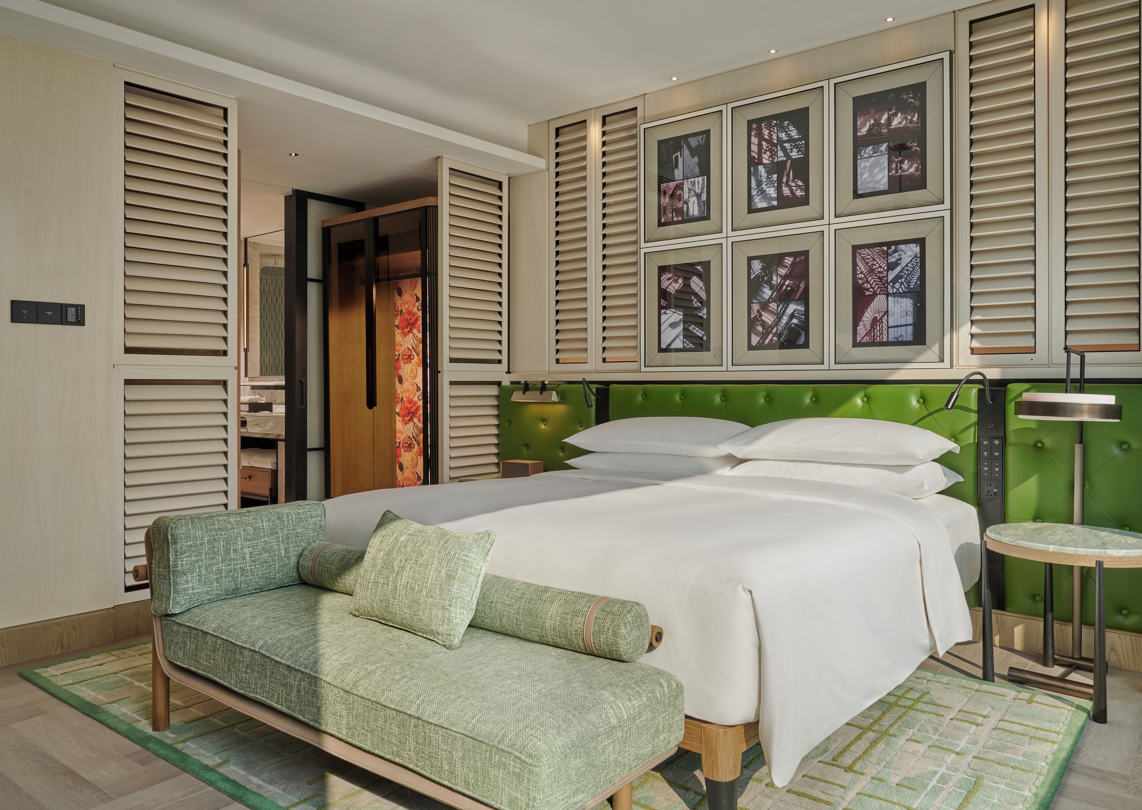 A modern hotel room with a large bed, a green sofa, and a green headboard. There are framed pictures on the wall and a small round table.