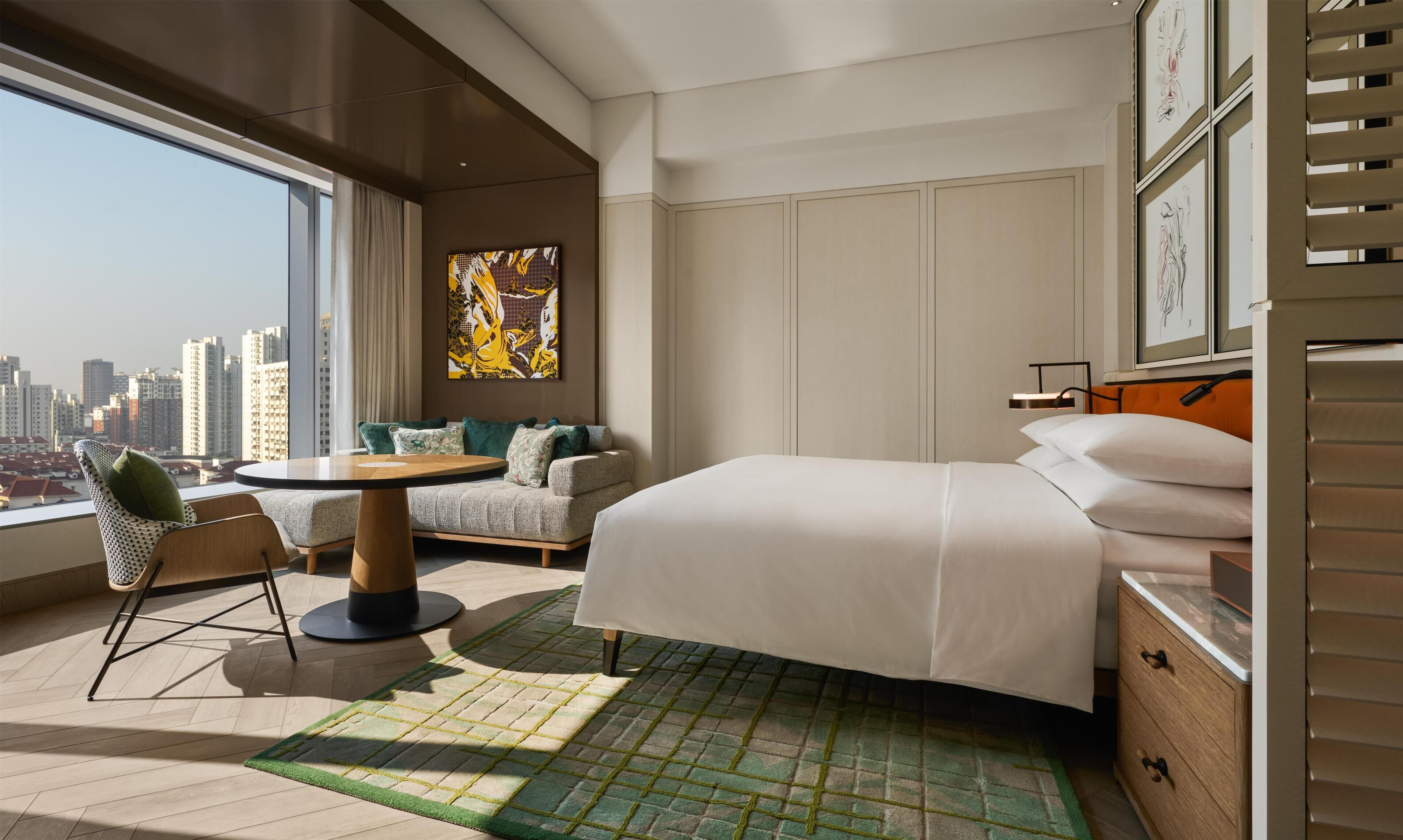 A hotel room with a large window, a bed, a seating area with a table, and artwork on the wall.