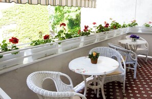 Une terrasse extérieure avec des meubles en osier et une table blanche, entourée d’une balustrade blanche agrémentée de plantes en pot.