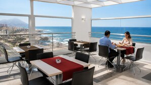 Un espace repas avec des tables et des chaises, de grandes fenêtres offrant une vue sur l’océan et le paysage urbain.