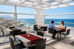 A dining area with tables and chairs, large windows with a view of the ocean and cityscape.