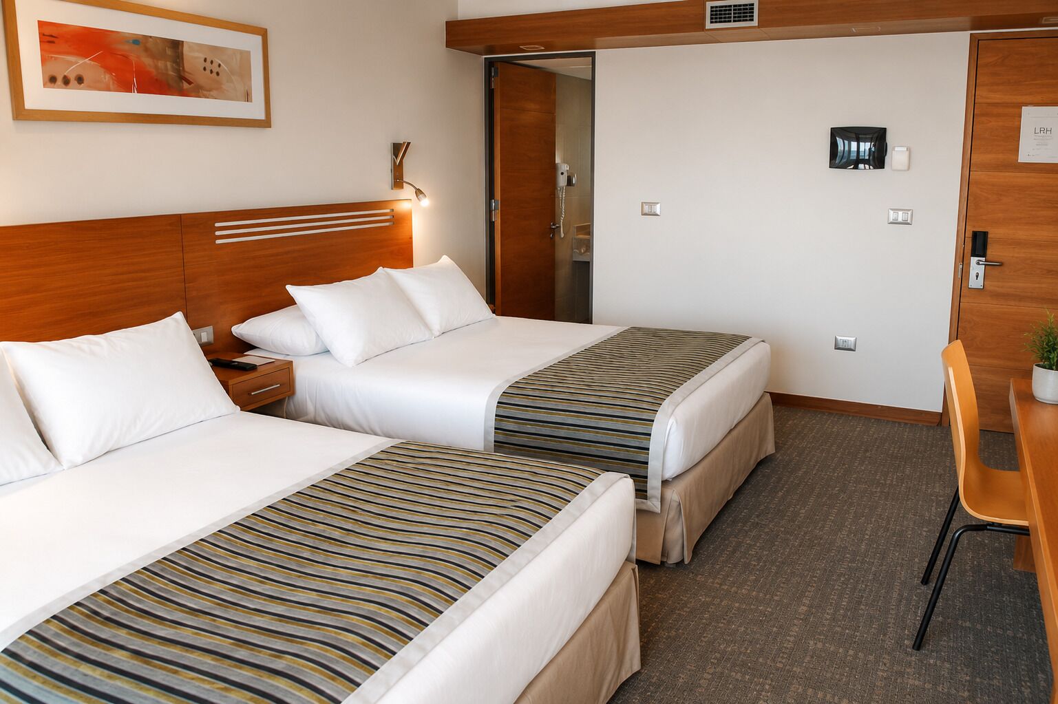 Une chambre d’hôtel avec deux lits, un bureau, une chaise, une table de chevet, une lampe et un tableau au mur.