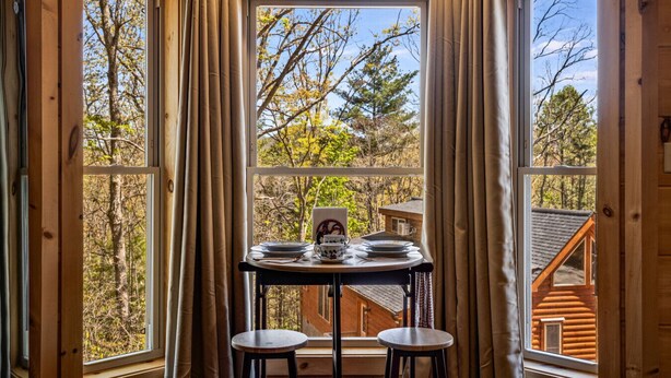 Eine Holzhütte mit Essbereich, inklusive Tisch und Stühlen, großen Fenstern mit Vorhängen sowie Blick auf Bäume und ein Gebäude.