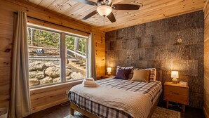 Ein Schlafzimmer einer Holzhütte mit einem Bett, Nachttischen, einem Deckenventilator und einem großen Fenster mit Blick auf eine felsige Landschaft.