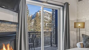 A room with a fireplace, a balcony with a stone wall, and a view of snow-capped mountains. - Amazing Views 1BR Condo w/ Hot Tub + Pool! (Canmore)