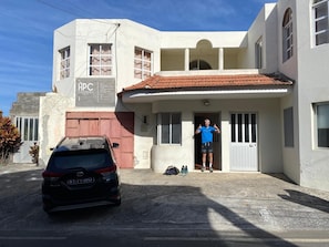 Un edificio de dos pisos con un coche estacionado frente a él, una persona de pie en la entrada, y un letrero con 'APC' en el lateral.