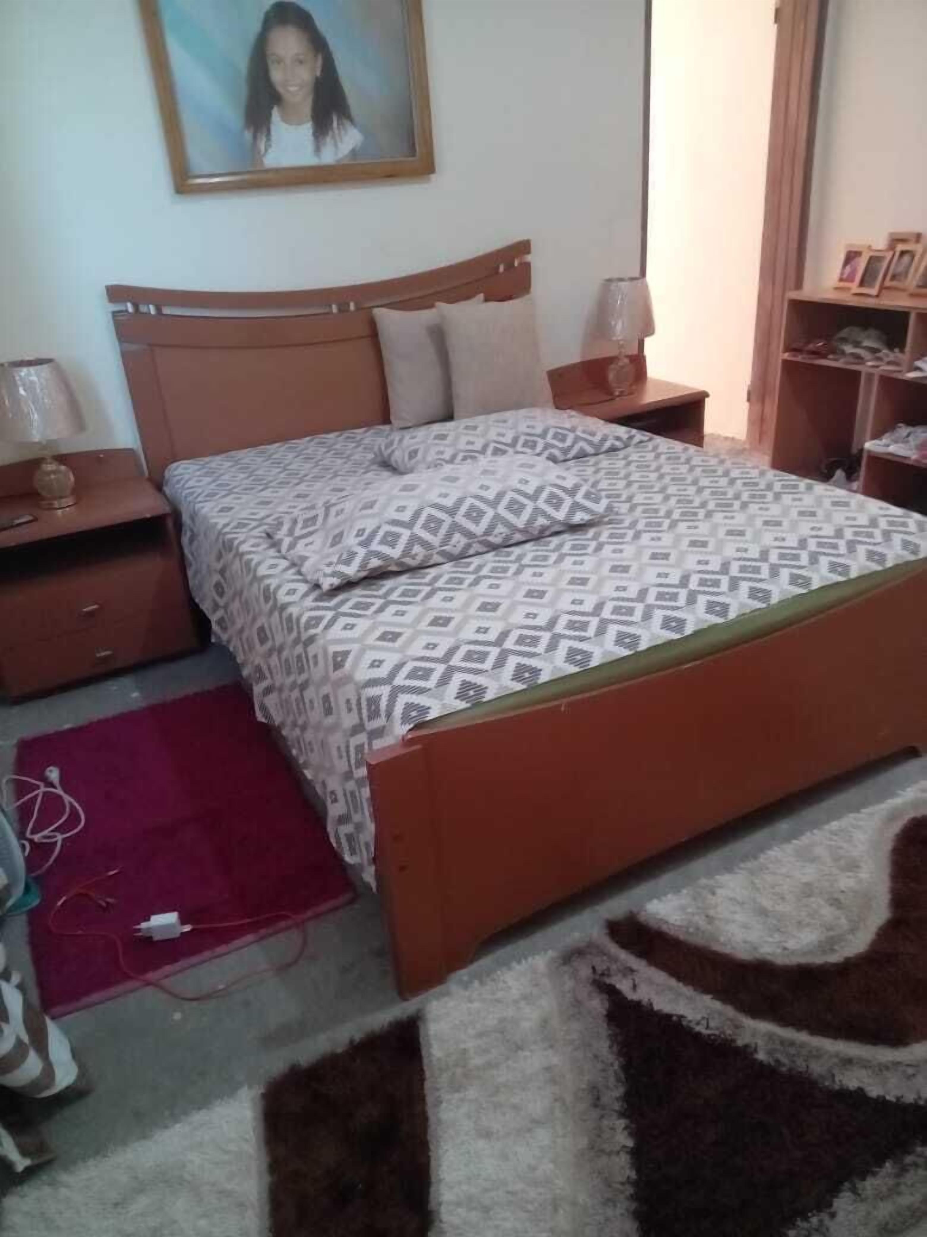 Un dormitorio con una cama de madera, dos mesitas de noche, un velador, una alfombra roja y un cuadro enmarcado en la pared.