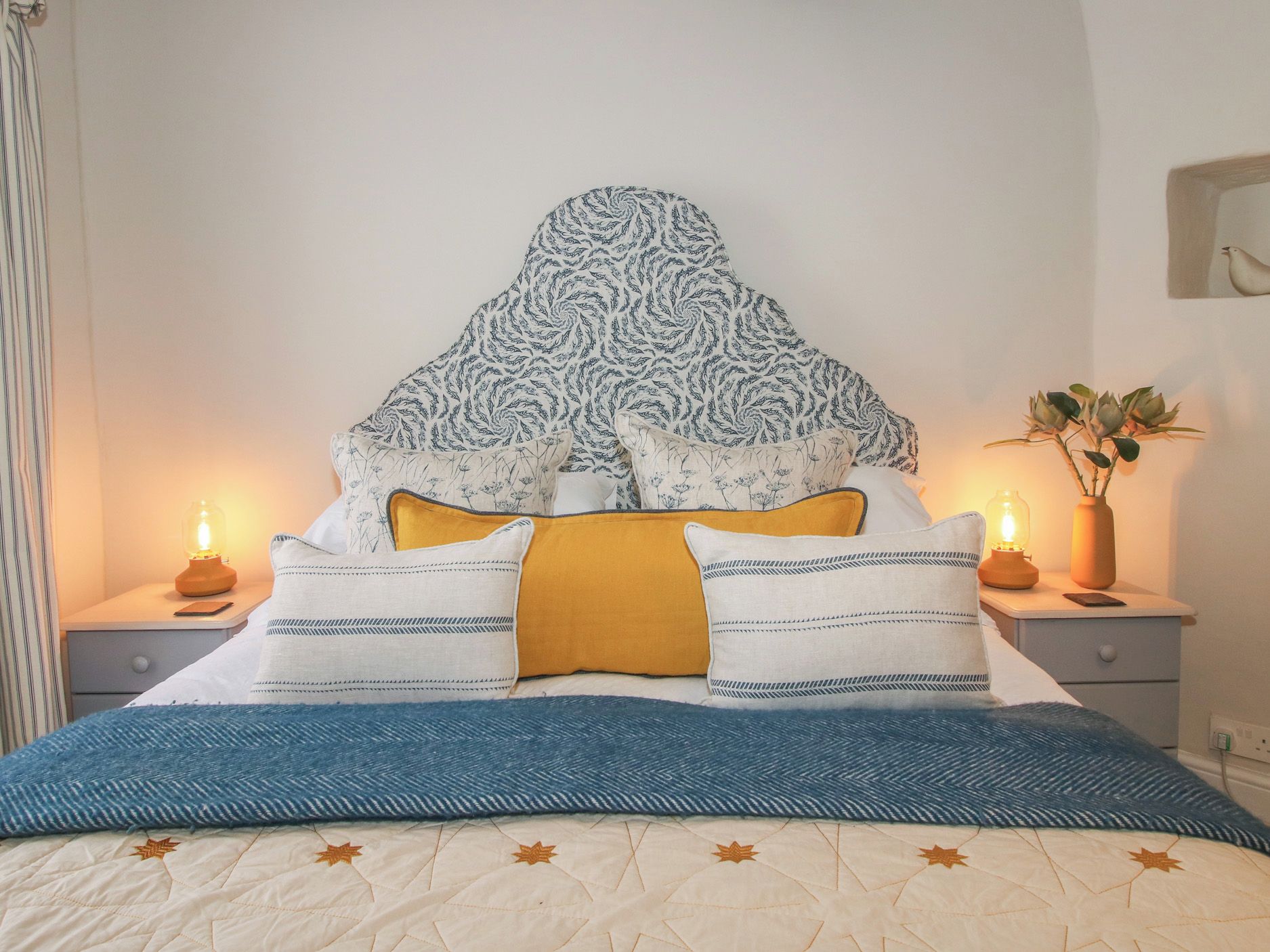 Una cama bien hecha con un cabecero estampado, varios almohadones y una colcha azul.