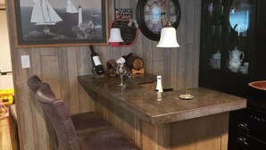 Uma área de bar com balcão de madeira, dois bancos altos, um quadro com barcos à vela, um relógio e uma placa de bar familiar.