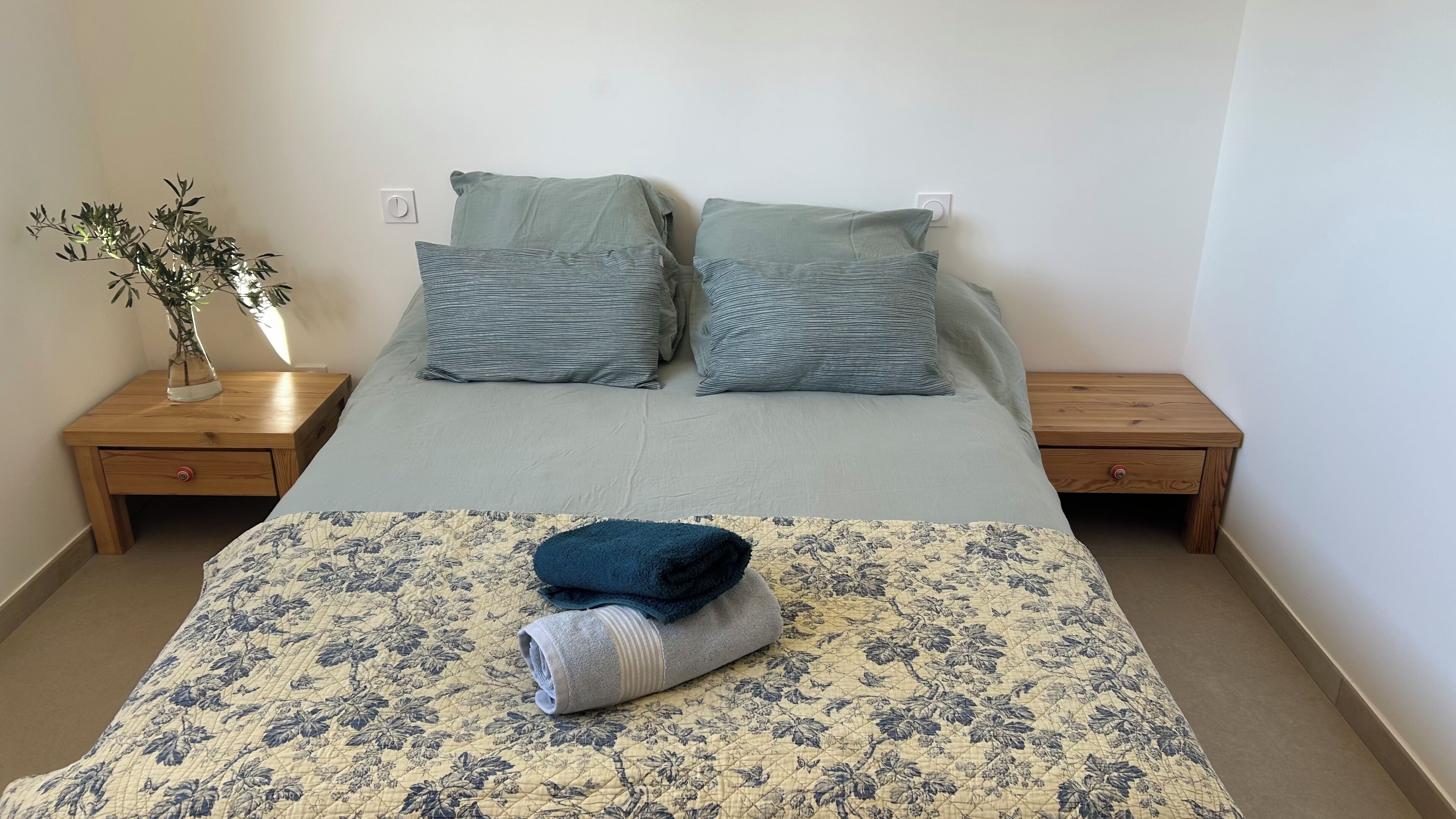 A bed with a floral quilt, two pillows, a vase with a plant, and two wooden nightstands.