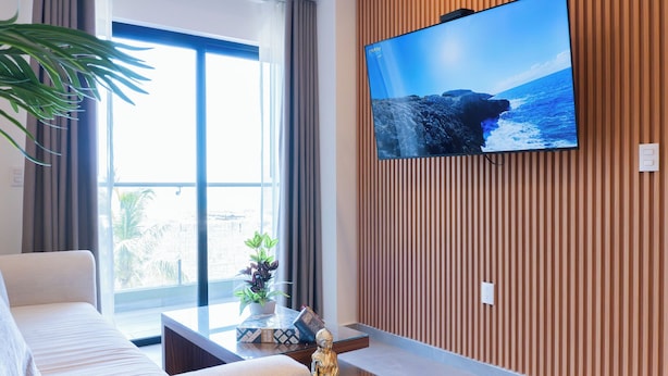 Un salon moderne doté d’un grand téléviseur à écran plat fixé à un mur en bois, d’un canapé, d’une table basse agrémentée d’objets décoratifs et offrant une vue sur l’extérieur grâce à de grandes fenêtres.