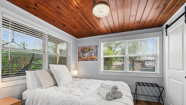 A bedroom with a wooden ceiling, white bedding, a painting on the wall, and a window with blinds.