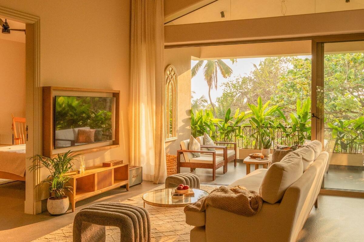 A spacious living area with a sofa set, a coffee table, and a television. There is a large window providing a view of lush greenery outside.