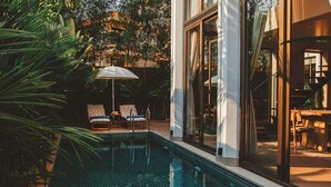 A modern outdoor pool area with two lounge chairs and a table, surrounded by lush greenery and large windows.