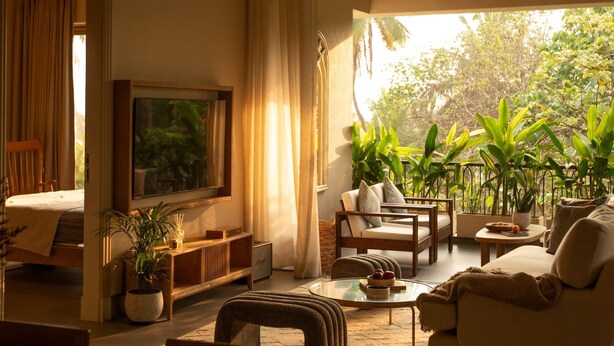 A cozy living room with a sofa, armchairs, a coffee table, and a TV. There are potted plants and a view of greenery outside.