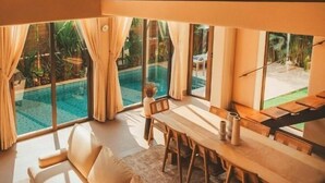 A spacious living area with a sofa, dining table, and chairs, large windows with curtains, and a view of a swimming pool.