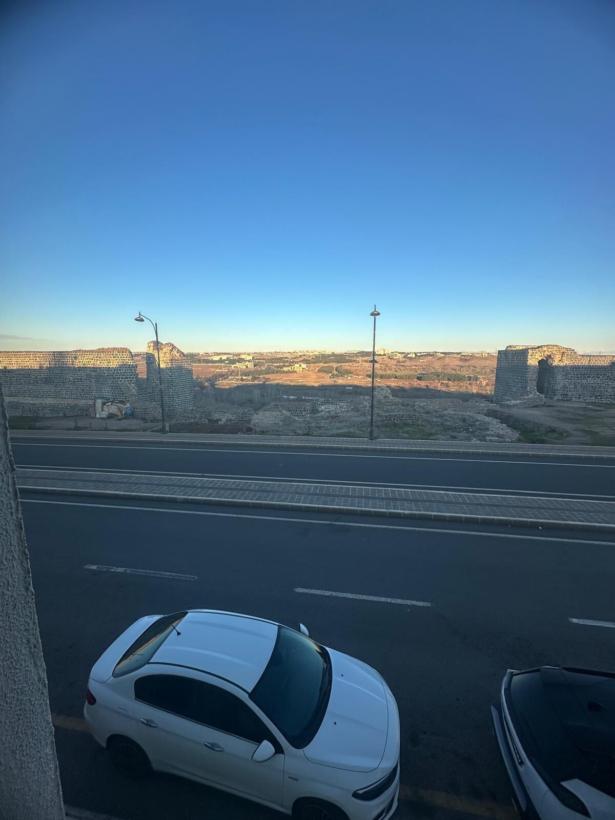 Et gadesyn med en hvid bil parkeret til siden, en klar blå himmel og et fjerntliggende vue af en stor, historisk murstensmur.