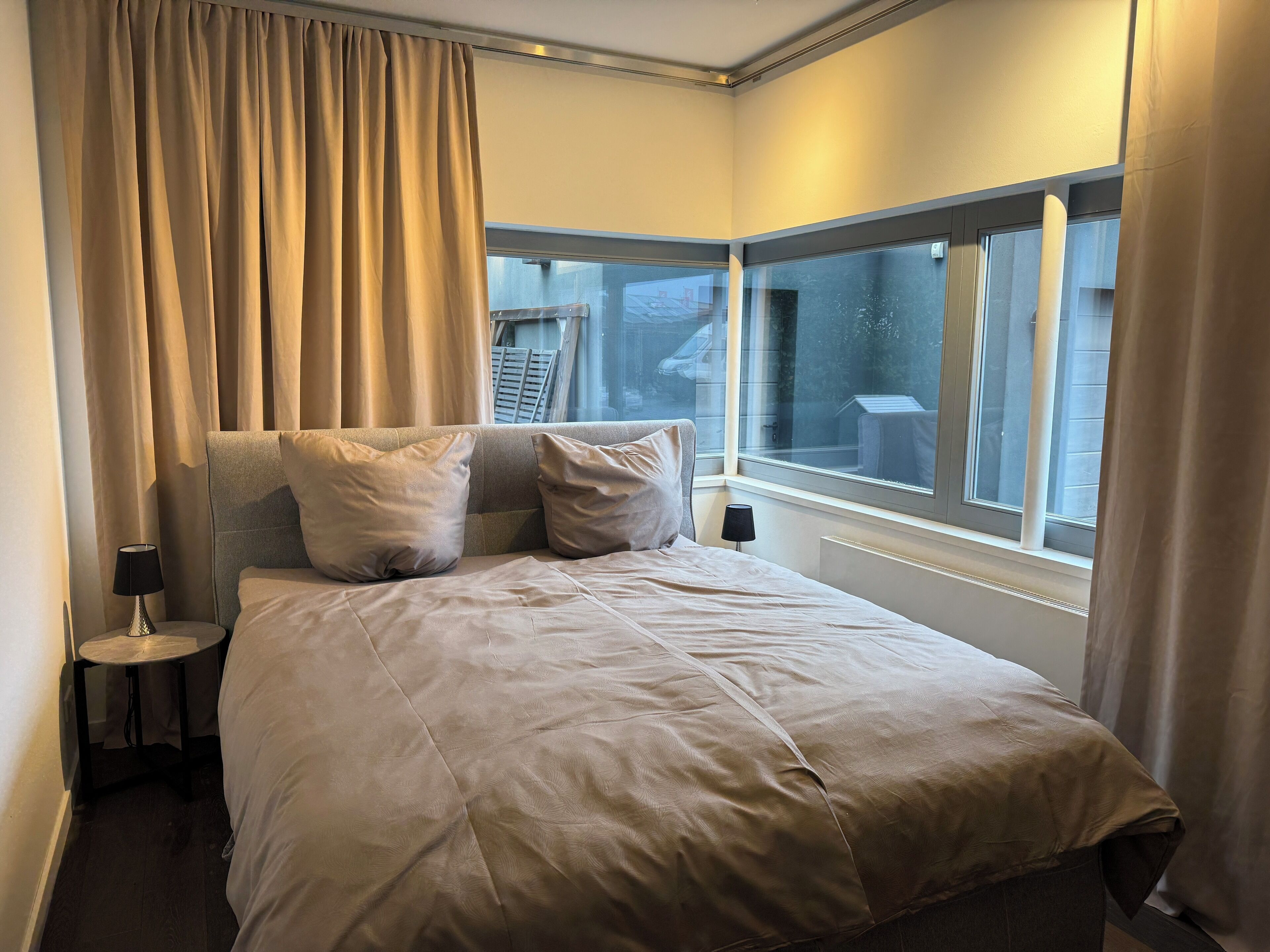 Ein ordentlich bezogenes Bett mit Kopfteil, zwei Kissen, einer Nachttischlampe und Blick durch ein mit Vorhängen versehenes Fenster nach draußen.