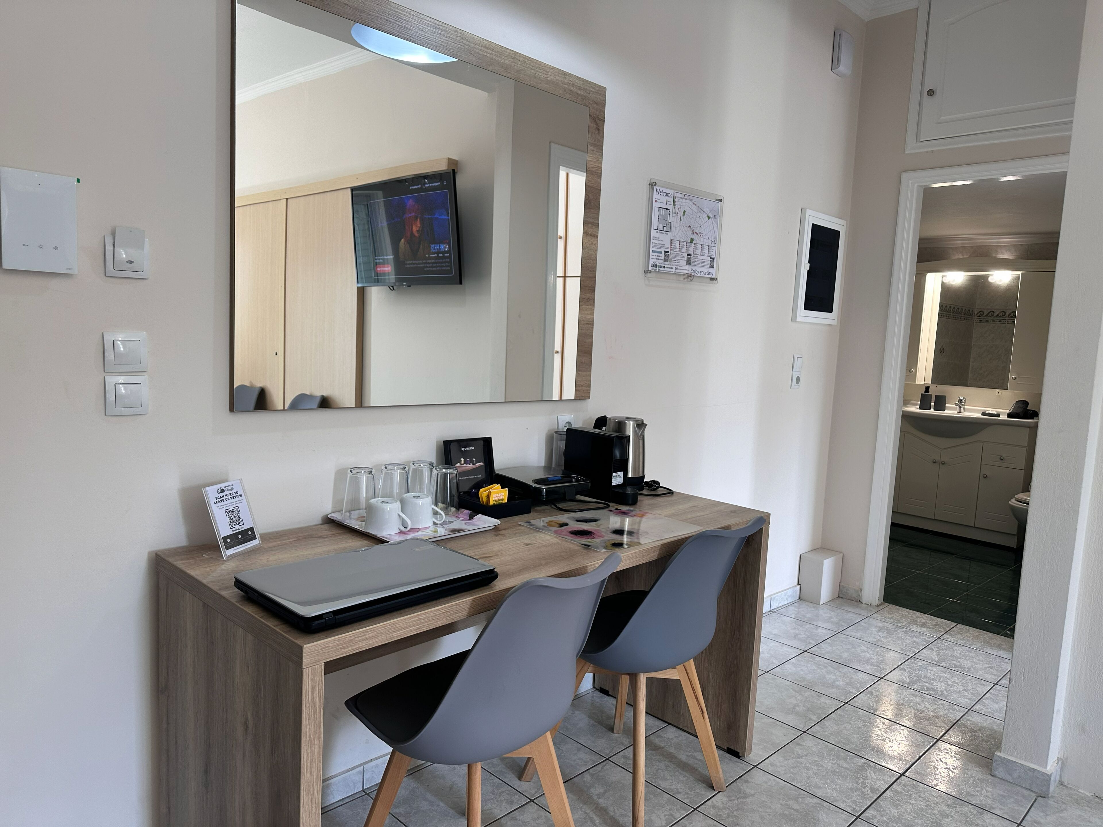 A modern hotel room with a wooden desk, two chairs, a laptop, a coffee maker, and a large mirror.