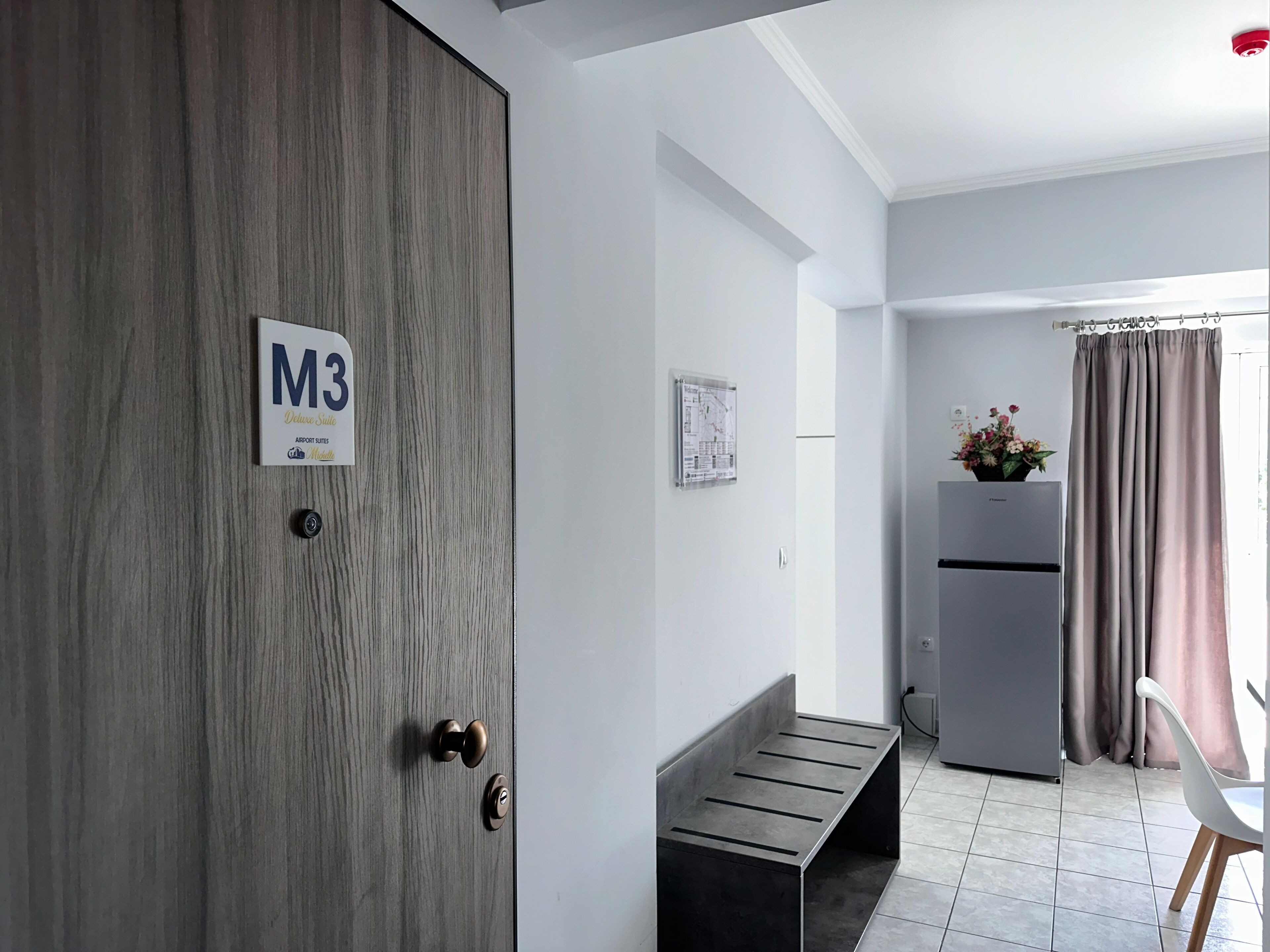 Ett modernt hotellrum med en trädörr märkt med ”M3”, en bänk, ett kylskåp och en matplats med en stol.