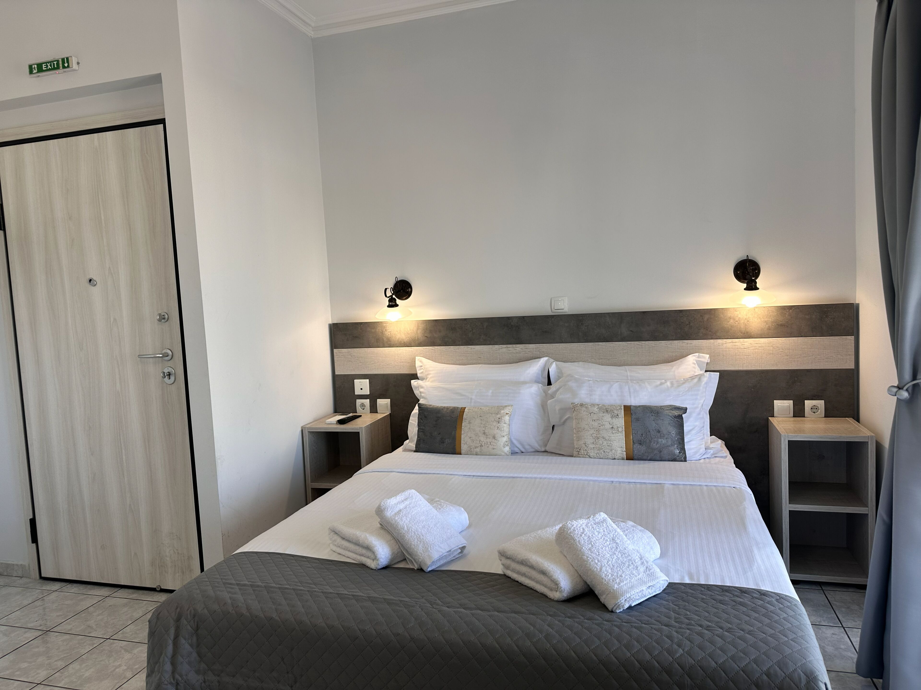 A hotel room with a bed, two bedside tables, a nightstand, a door, and a wall-mounted light fixture.