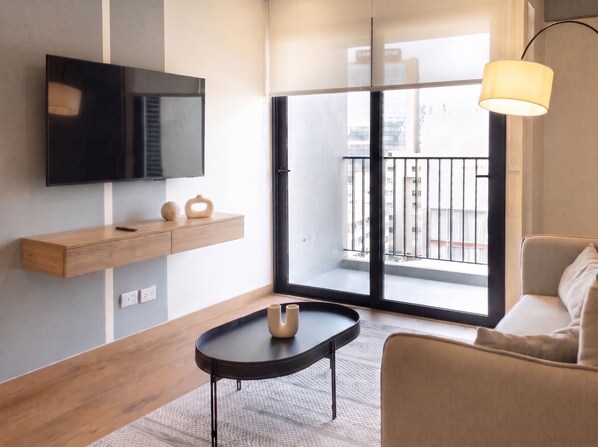 A modern living room with a flat-screen TV, a wooden shelf, a round coffee table, a sofa, and a balcony with a view. - w* | Charming 2BR w/ Balcony in Miraflores (Miraflores)