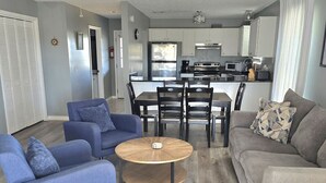 Ein modernes Wohnzimmer mit einem runden Couchtisch, blauen Sesseln und einem grauen Sofa. Im Hintergrund sind ein Deckenventilator und eine Küche zu sehen.