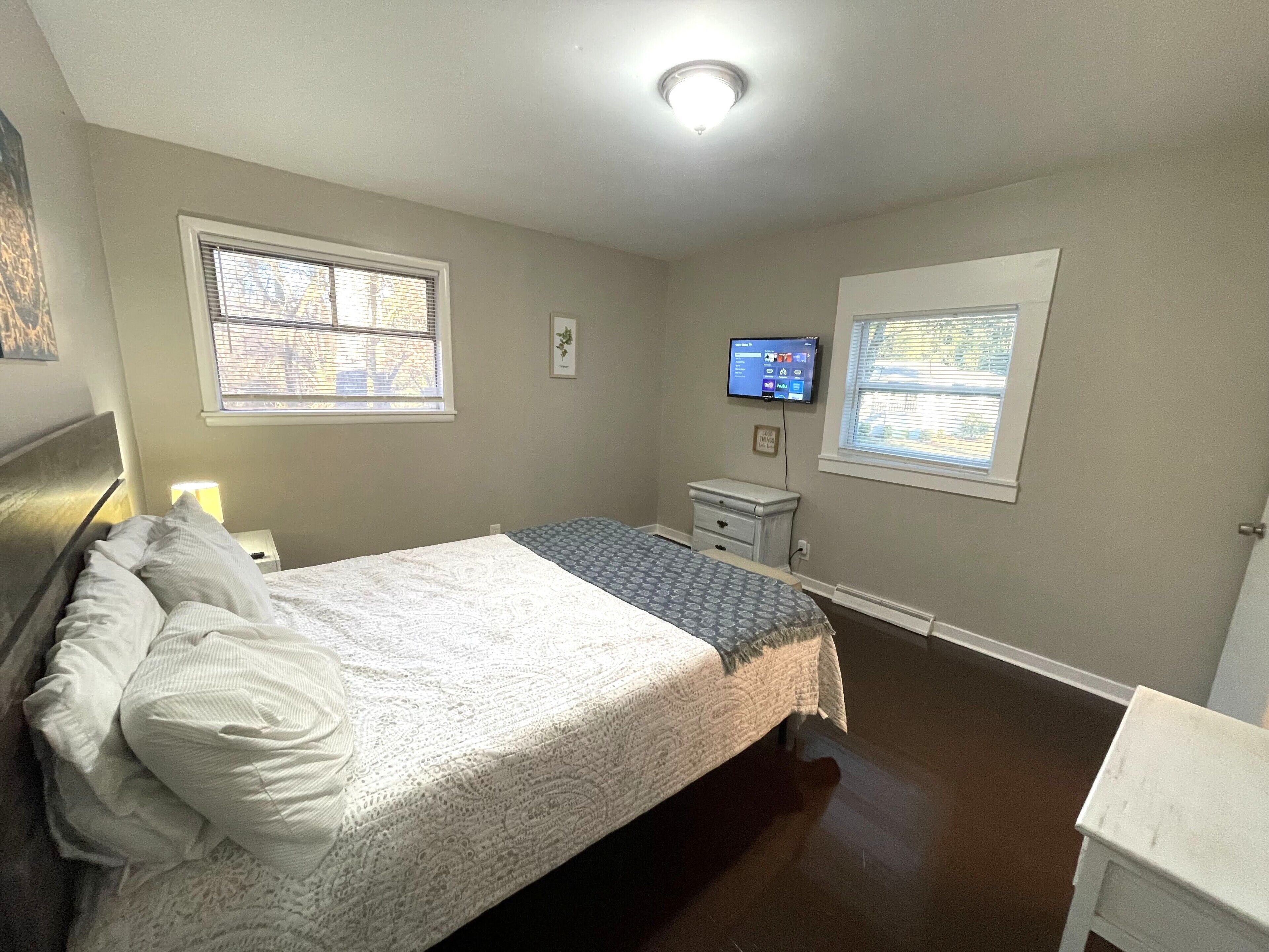 A bedroom with a bed, nightstand, dresser, TV, and two windows.