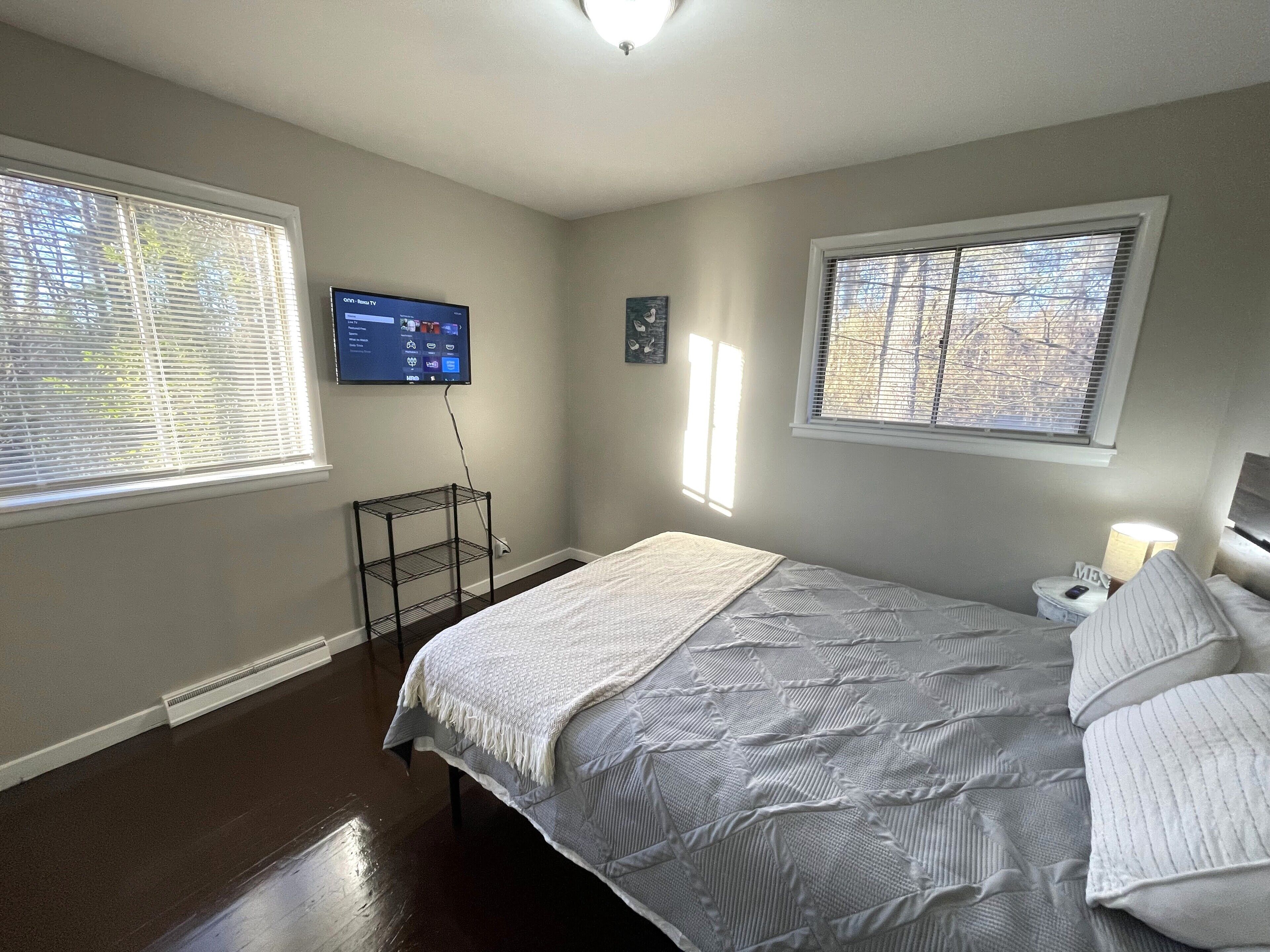 Un dormitorio con cama, televisión montada en la pared y dos ventanas con persianas.