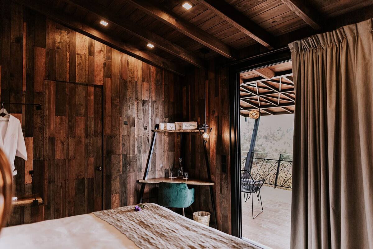 A rustic wooden room with a bed, a hanging light fixture, and a view of an outdoor area with a geometric structure.