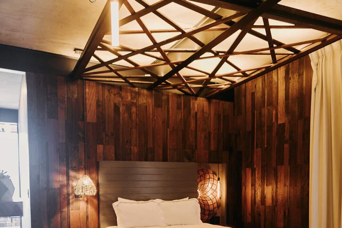 A bedroom with a wooden ceiling, a bed with white bedding, a round wooden table, and two wall-mounted lamps.