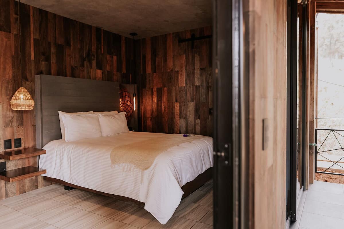 A bedroom with a wooden wall panel design, a bed with white bedding, a bedside table with a lamp, and a view through an open door.