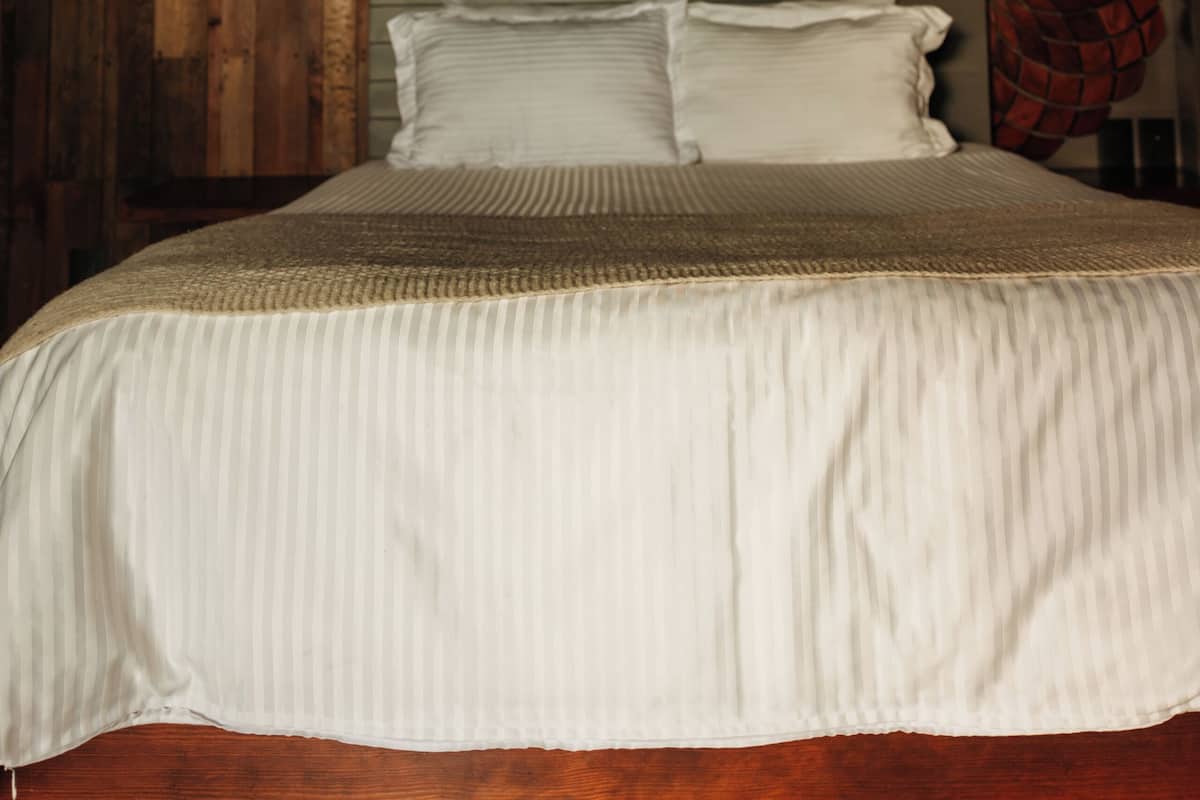 A bed with a wooden headboard, a textured white bedspread, and a woven lamp on the left.