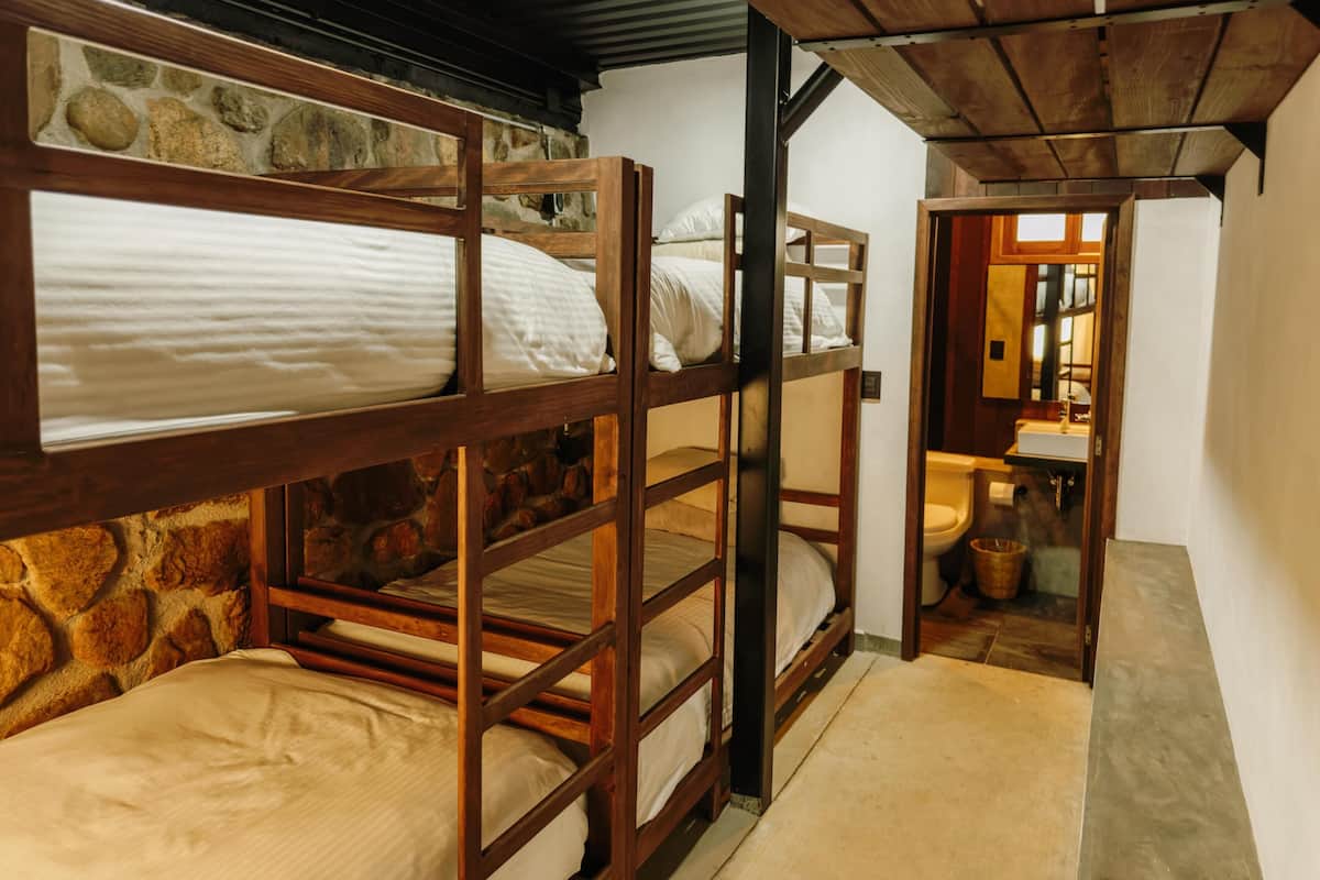 A bunk bed room with stone walls, a wooden ceiling, and a visible bathroom with a sink and toilet.