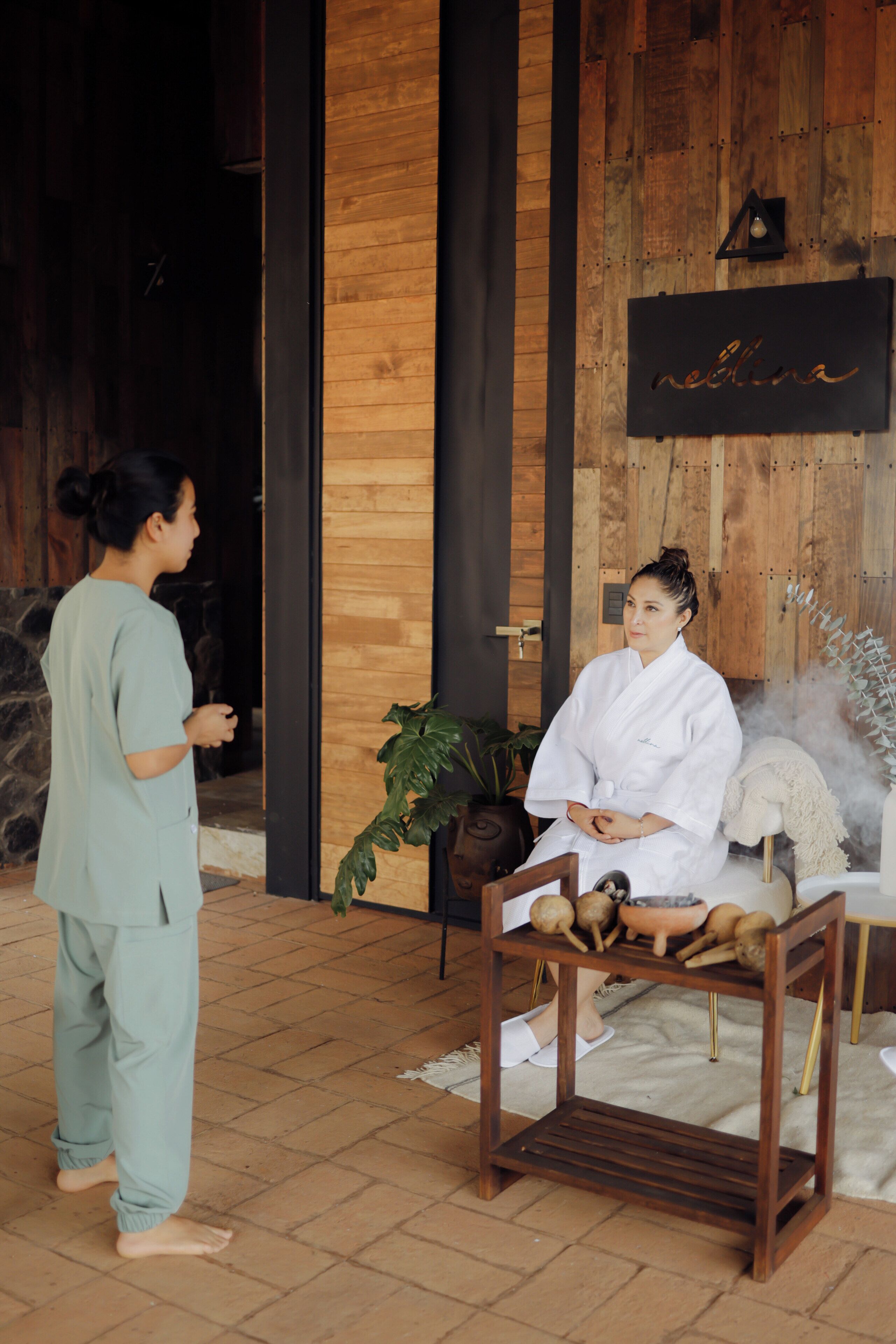 A spa setting with a wooden wall, a sign with the word "neblina," and a person in a bathrobe sitting on a wooden bench.