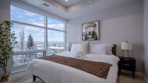 A bedroom with a large window, a bed, a nightstand, and a potted plant.