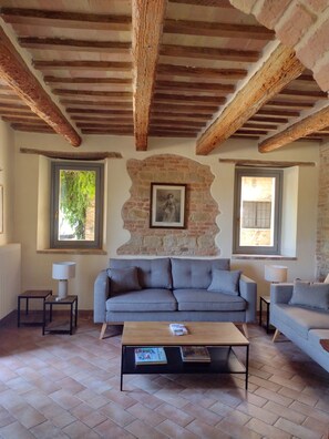 A cozy living room with a grey sofa, a wooden coffee table, and two side tables. A stone wall with a picture frame is behind the sofa. - Casa Alta - 4 Bedroom Stunning Views and Villa with Pool in Medieval  Hill Town (Sant'Angelo in Pontano)